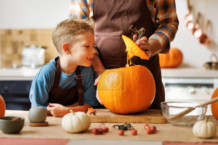 Photo for Cute happy little boy helping his father to carve Halloween pumpkin while standing in kitchen at home and preparing for autumn holiday, family son and dad making Jack-o-Lantern together - Royalty Free Image