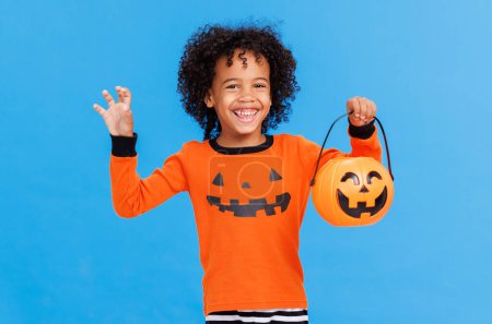 Foto de Feliz niño afroamericano alegre con el pelo rizado en traje de calabaza gestos de miedo y celebra Halloween y se ríe en fondo azul brillante - Imagen libre de derechos