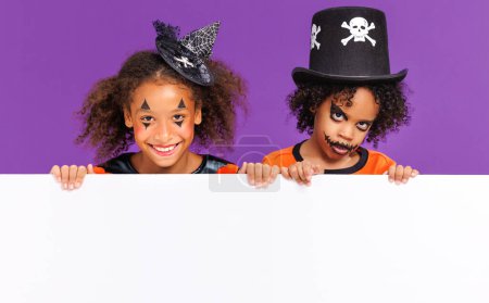 Photo for Happy Halloween! African american children in carnival costumes and makeup with blank white poster on colored purple background - Royalty Free Image