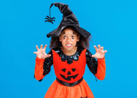 Téléchargez les photos : Heureuse fille afro-américaine joyeuse avec des cheveux bouclés en costume de citrouille gestes effrayants et célèbre Halloween et rit sur fond bleu vif - en image libre de droit
