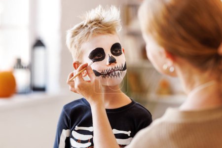 Festliches Make-up für Halloween. Frau beim Skelett-Make-up für einen kleinen fröhlichen Jungen im Kostüm, während sie den Urlaub zu Hause vorbereitet.