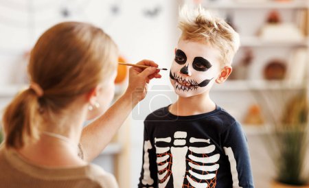 Téléchargez les photos : Maquillage festif pour Halloween. Femme faisant squelette maquillage pour un petit garçon joyeux en costume tout en préparant des vacances à la maison. - en image libre de droit