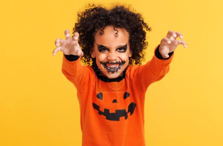 Téléchargez les photos : Joyeux garçon afro-américain joyeux avec les cheveux bouclés en costume de citrouille et terrible maquillage gestes effrayants et célèbre Halloween et rit sur fond jaune vif - en image libre de droit