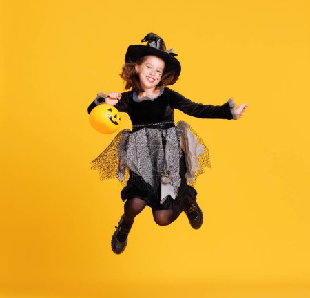 Téléchargez les photos : Joyeux gai fille en costume de sorcière avec panier de citrouille sautant et célèbre Halloween et rit sur fond jaune - en image libre de droit