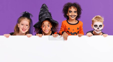 Téléchargez les photos : Joyeux Halloween ! Groupe d'enfants en costumes de carnaval et maquillage avec affiche blanche vierge sur fond violet coloré - en image libre de droit