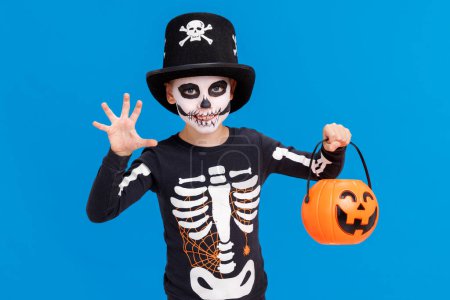 Foto de Feliz niño alegre en traje de esqueleto con cesta de calabaza celebra Halloween y gesto de miedo sobre fondo azul brillante - Imagen libre de derechos