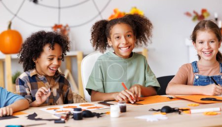 Photo for Happy smiling multinational group of children  making Halloween home decorations together, kids painting pumpkins and making paper cuttings - Royalty Free Image