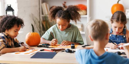 Photo for Happy smiling multinational group of children  making Halloween home decorations together, kids painting pumpkins and making paper cuttings - Royalty Free Image