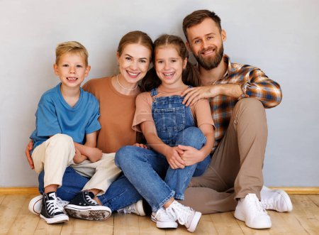 Photo for Happy family mother father and children sitting on floor against empty grey wall  in new apartment after moving - Royalty Free Image