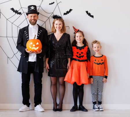 Téléchargez les photos : Mère de famille joyeuse père et enfants en costumes de carnaval célèbrent Halloween près du mur gris avec des toiles d'araignée et des chauves-souris - en image libre de droit
