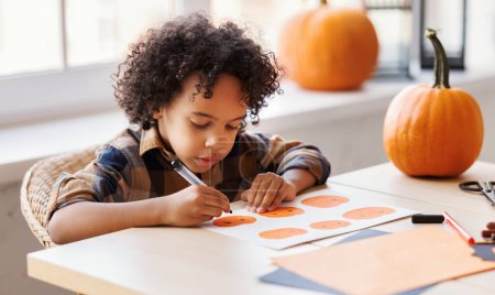Photo for African american cute kid boy  making Halloween home decorations  while sitting at wooden table, child painting pumpkins and making paper cuttings - Royalty Free Image