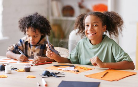 Photo for Happy african american children girl and boy making Halloween home decorations together, kids painting pumpkins and making paper cuttings - Royalty Free Image