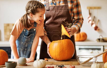 Photo for Cute happy girl helping father to carve Halloween pumpkin while standing in kitchen at home and preparing for autumn holiday, family daughter and dad making Jack-o-Lantern together - Royalty Free Image