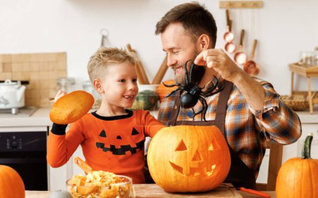 Photo for Cute happy   boy helping his father to carve Halloween pumpkin, playing with spider and preparing for autumn holiday, family son and dad making Jack-o-Lantern together - Royalty Free Image