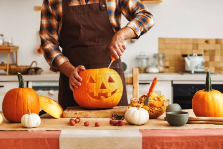 Photo for Cropped shot of  man in apron standing in kitchen and carving large orange pumpkin for Halloween party while  making scary face on jack-o-lantern with knife to set the mood for trick-or-treaters - Royalty Free Image