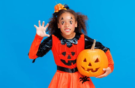 Photo for Happy cheerful african american girl with  jack-o-lantern   in  pumpkin Halloween costume scary gestures on  blue background - Royalty Free Image