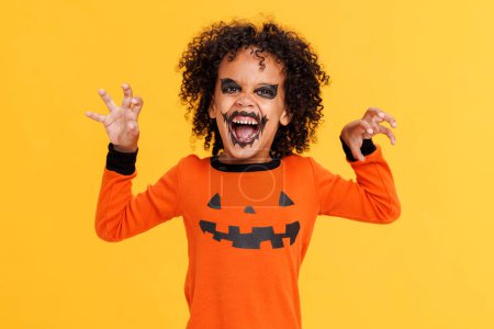 Foto de Feliz niño afroamericano alegre con el pelo rizado en traje de calabaza y maquillaje terrible hacer gestos de miedo y celebra Halloween y se ríe en el fondo amarillo - Imagen libre de derechos