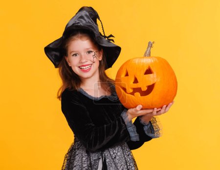 Téléchargez les photos : Joyeux gai fille en costume de sorcière avec citrouille Jack-o-lanterne célèbre Halloween et rit sur fond jaune vif - en image libre de droit