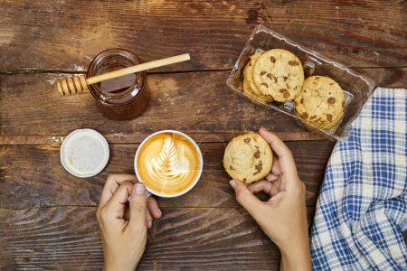 Photo for Woman take her breakfast, Latte art on hot latte coffee with homemade chocolate chip cookies with honey jar on wood table - Royalty Free Image
