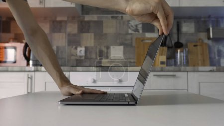 Konzept der Arbeit zu Hause während der Isolation. Seitenansicht männlicher Hände beim Öffnen eines neuen silbernen Laptops, der auf dem weißen Tisch in der Küche steht.