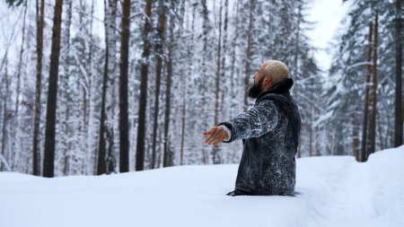 Glücklicher junger Mann im märchenhaften Winterwald, der auf Knien zu Gott betet. Medien. Konzept der Religion und Gefühl der Einheit mit der Natur