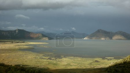 Sun and shadow on green meadow and a lake. Creative. Calm water and mountains on cloudy sky background