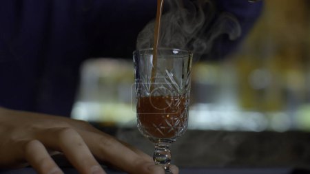 Close up of bartender at a bar counter pouring alcohol into vintage glass. Media. Pouring red hot alcoholic tincture with steam