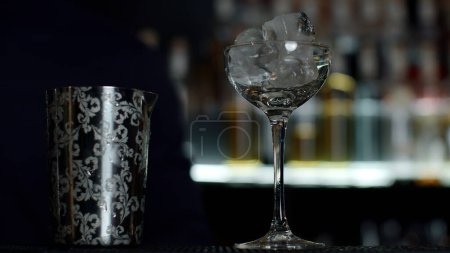 Close up of a bartender putting ice cubes into the small empty cocktail glass. Media. Details of making a drink with blurred bottles of alcohol on the background