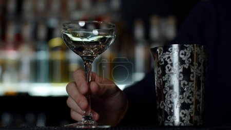 Großaufnahme eines Barkeepers, der Eiswürfel in das kleine leere Cocktailglas gibt. Medien. Details der Herstellung eines Getränks mit verschwommenen Flaschen Alkohol auf dem Hintergrund