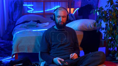 Young bearded blond man sitting on the floor by his bed. Media. Man shaking head while listening music in bedroom with neon light lamps