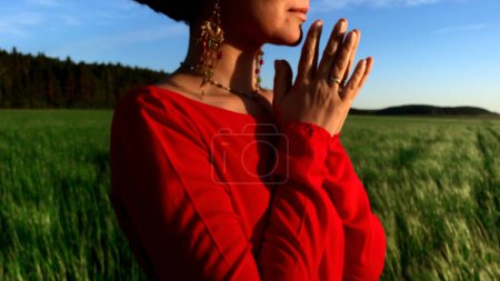 Mujer bonita joven meditando con los ojos cerrados. Clip de stock. Orando mientras está de pie en un campo con una hermosa vista en un día de verano ventoso al atardecer