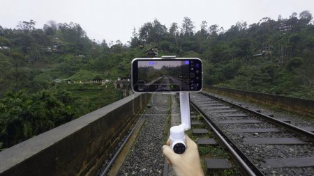 Auf Reisen mit dem Stativ fotografieren. Handeln. Videoaufnahmen am Telefon mit Stabilisator während der Reise. Schießen Brücke mit Eisenbahn im Dschungel am Telefon für Blog.