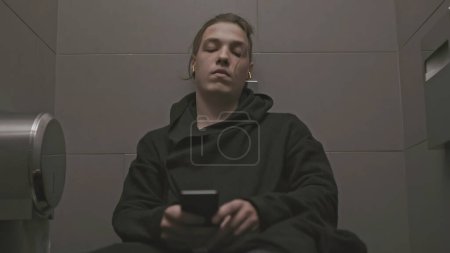 Jeune homme avec dépression dans les toilettes. Des actions. L'homme s'assoit sur les toilettes et écoute de la musique triste. Adolescent caché dans la cabine de toilette et est triste.