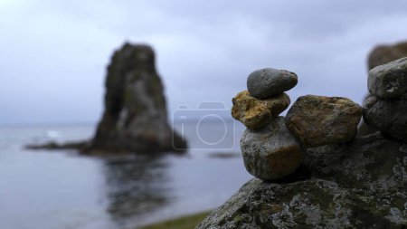 Close-up of stacked rocks on beach with rocks. Clip. Stones stacked in composition on blurred background of rocks in sea. Stones stacked in turret stand on background of overcast sea.