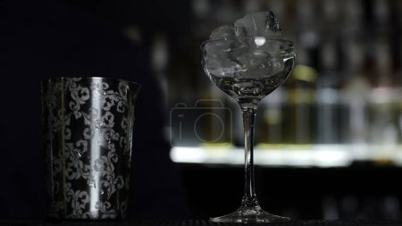 Großaufnahme eines Barkeepers, der Eiswürfel in das kleine leere Cocktailglas gibt. Medien. Details der Herstellung eines Getränks mit verschwommenen Flaschen Alkohol auf dem Hintergrund
