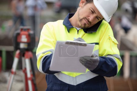 surveyor on site writing on clipboard and talking on smartphone
