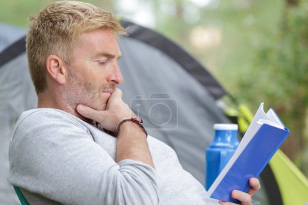 middle-aged man engrossed in book by his tent