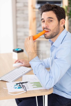 young man at computer desk eating raw carrot