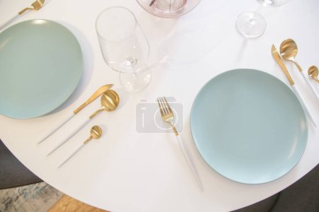 Foto de Mesa de comedor con platos de cerámica azul, cubiertos de oro con asas blancas y copas de vino, vista superior. - Imagen libre de derechos