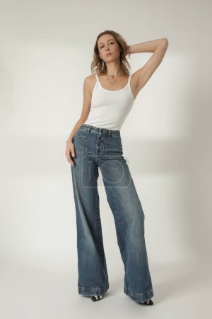 Téléchargez les photos : Portrait de mode de jeune femme en débardeur blanc et large jambe jeans bleu sur le fond blanc - en image libre de droit