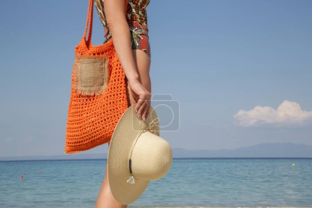 Photo for Young woman in tropical printed swimwear and straw hat cary orange crochet tote bag empty sandy beach on, summer fashion style, relaxed vacation - Royalty Free Image