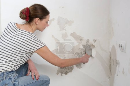 Salpeter auf dem Wandproblem. Frau kratzt und entfernt lose Farbe und schlechten Putz mit einem Schaber, bis eine feste Oberfläche erreicht ist. 