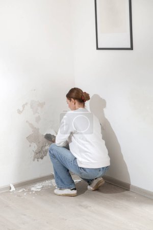 Saltpeter on the wall problem. Woman is using a scraper to scrape and remove all loose paint and plaster that is in poor condition, until a firm surface is achieved. 