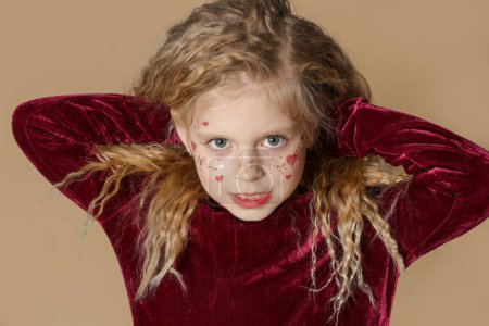 Photo for Portrait of cute little girl in red dress with long blonde hair and glitters and red hearts on the face - Royalty Free Image