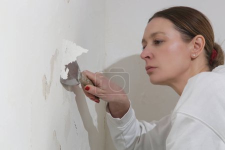 Salpeter auf dem Wandproblem. Frau kratzt und entfernt lose Farbe und schlechten Putz mit einem Schaber, bis eine feste Oberfläche erreicht ist.