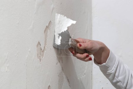 Salitre en el problema de pared. La mujer está usando un rascador para raspar y eliminar toda la pintura suelta y el yeso que está en mal estado, hasta que se logra una superficie firme.
