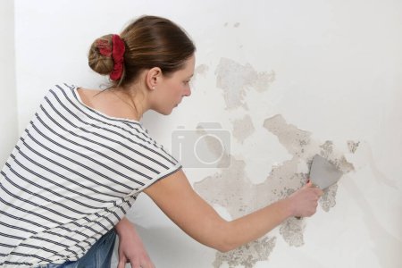 Salpeter auf dem Wandproblem. Frau kratzt und entfernt lose Farbe und schlechten Putz mit einem Schaber, bis eine feste Oberfläche erreicht ist.