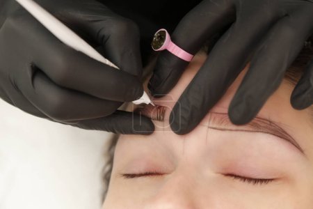 Microblading artist in a black gloves mixing pigment in ink ring cup. Close-up of pigment ring container and ink for eyebrow tattoo. Beauty procedure preparation.
