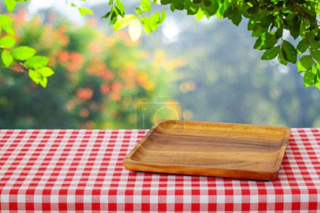Bandeja de madera vacía en la mesa con mantel rojo sobre desenfoque árboles con fondo bokeh, para montaje de alimentos y exposición de productos