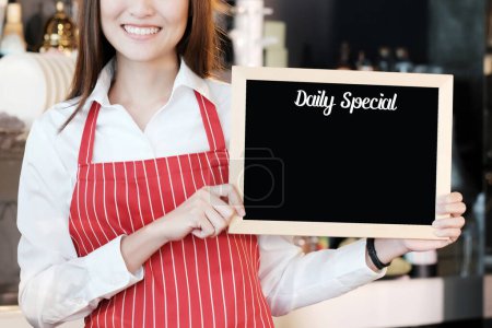 Smiling woman holding blank daily special board over blur cafe background, copy space for text, food and drinks background